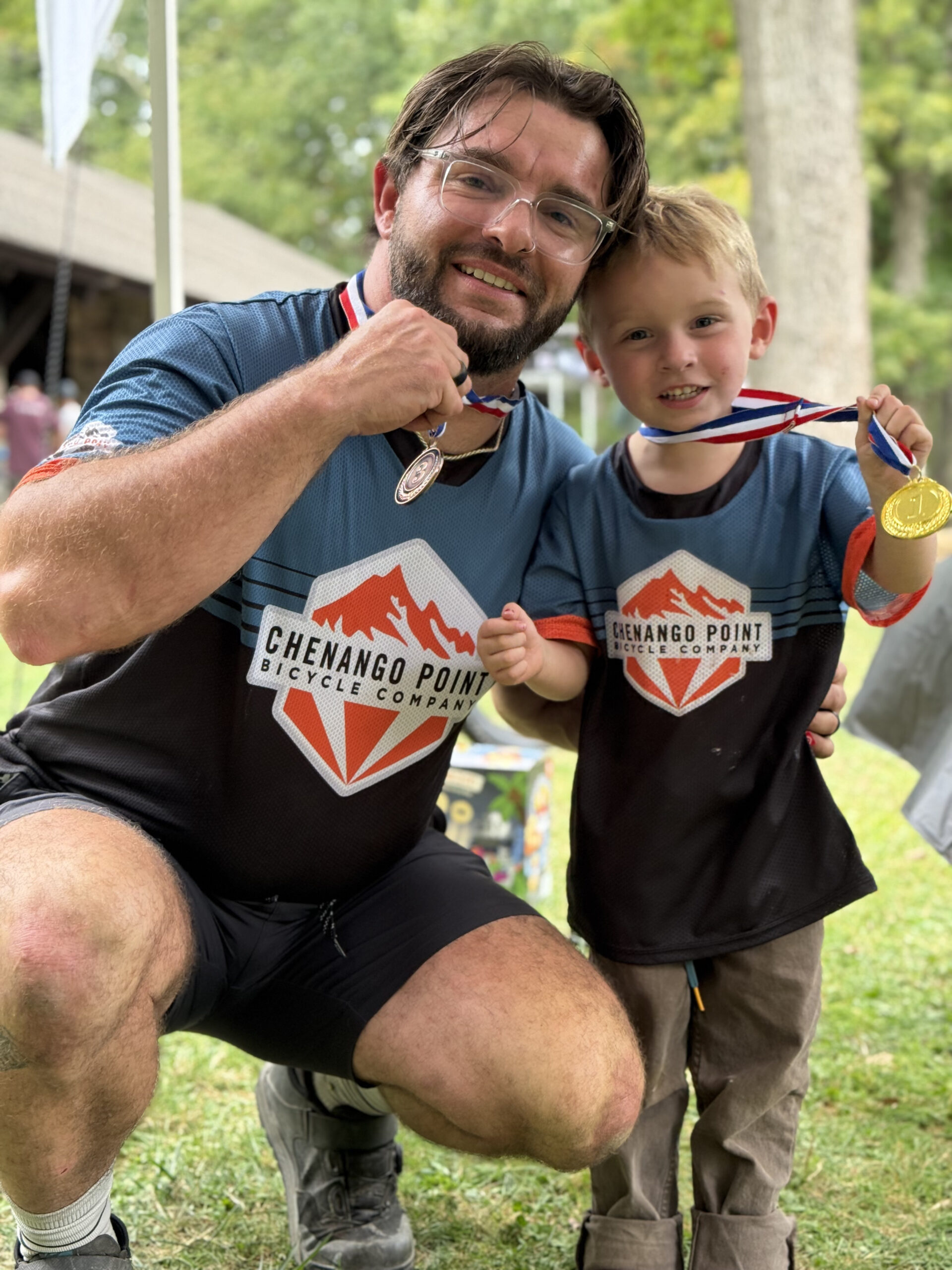 A dad and his young son wearing Chenango Point Bicycle Company jerseys, both holding up medals after an event